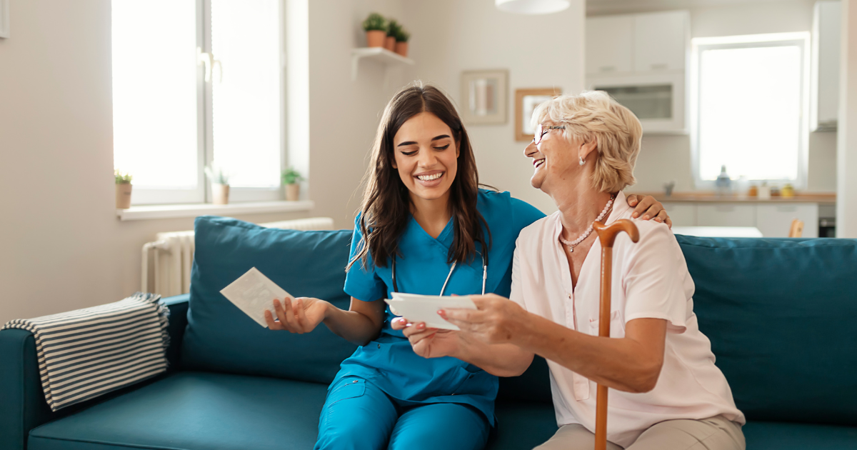 A smiling caregiver in blue scrubs sits beside an elderly woman on a couch while helping her review paperwork in a bright living room.