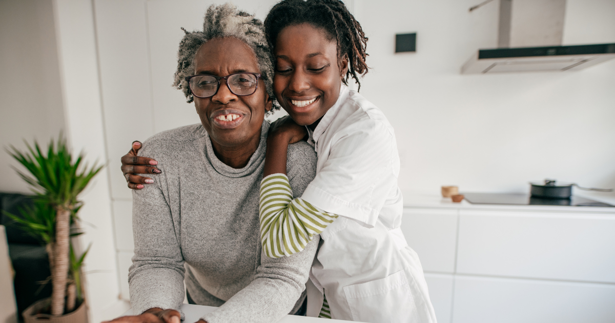 Smiling professional caregiver hugging an elderly woman in a bright kitchen, showing warmth, comfort, and in-home senior care support.