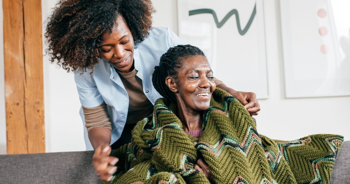 A professional caregiver gently adjusts a blanket around a smiling senior woman seated on a couch, providing warmth, comfort, and emotional support during an in-home care visit.