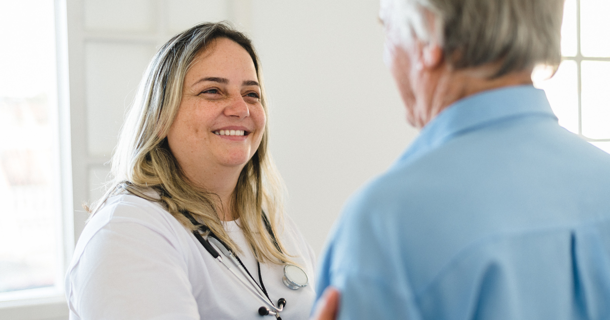 A compassionate home health nurse with a stethoscope smiles while speaking with a senior man in a bright home setting during an in-home care visit.