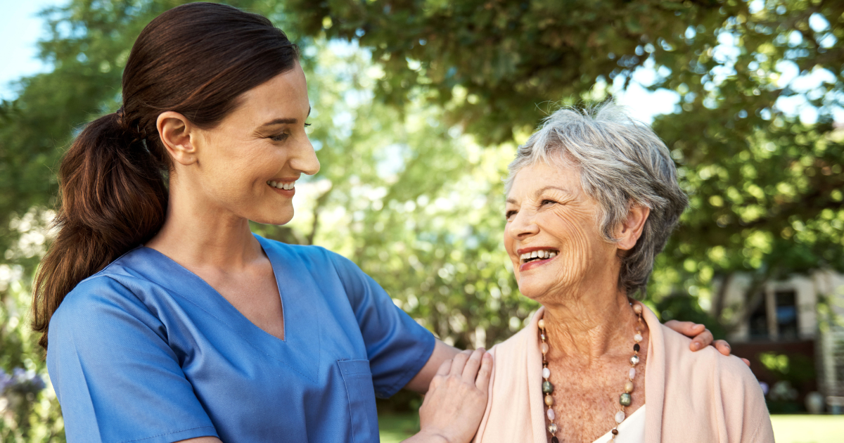 Smiling caregiver standing beside an elderly woman outdoors, offering supportive companionship in a garden setting.