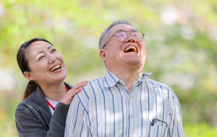 Smiling caregiver standing beside an elderly man as they laugh together during an outdoor companionship care visit.