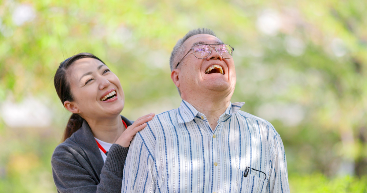 Smiling caregiver standing beside an elderly man as they laugh together during an outdoor companionship care visit.