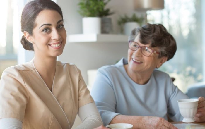 Caregiver sitting beside an elderly woman at a table as they share a warm conversation and enjoy tea together.
