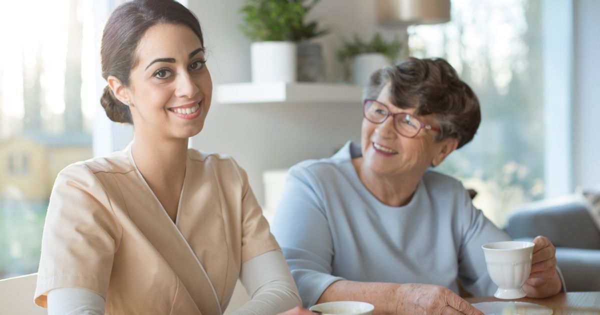 Caregiver sitting beside an elderly woman at a table as they share a warm conversation and enjoy tea together.