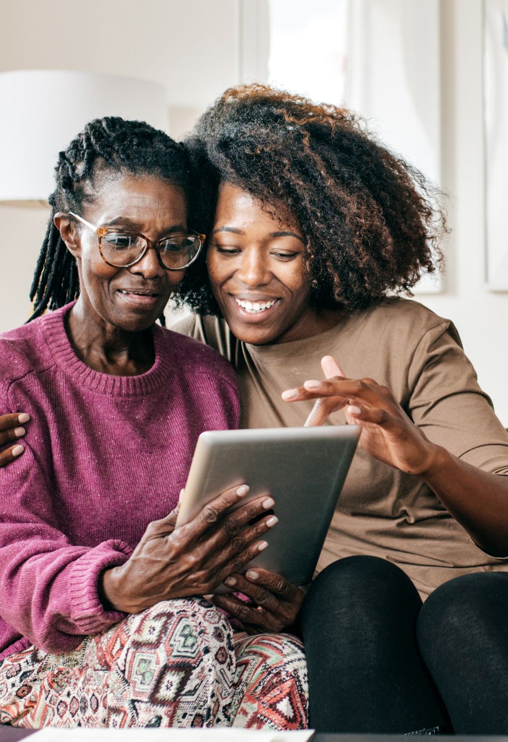Multi-Generational Women Using Tablet A young Black woman smiling and pointing at a tablet screen she is holding with an older Black woman wearing glasses and a purple sweater.