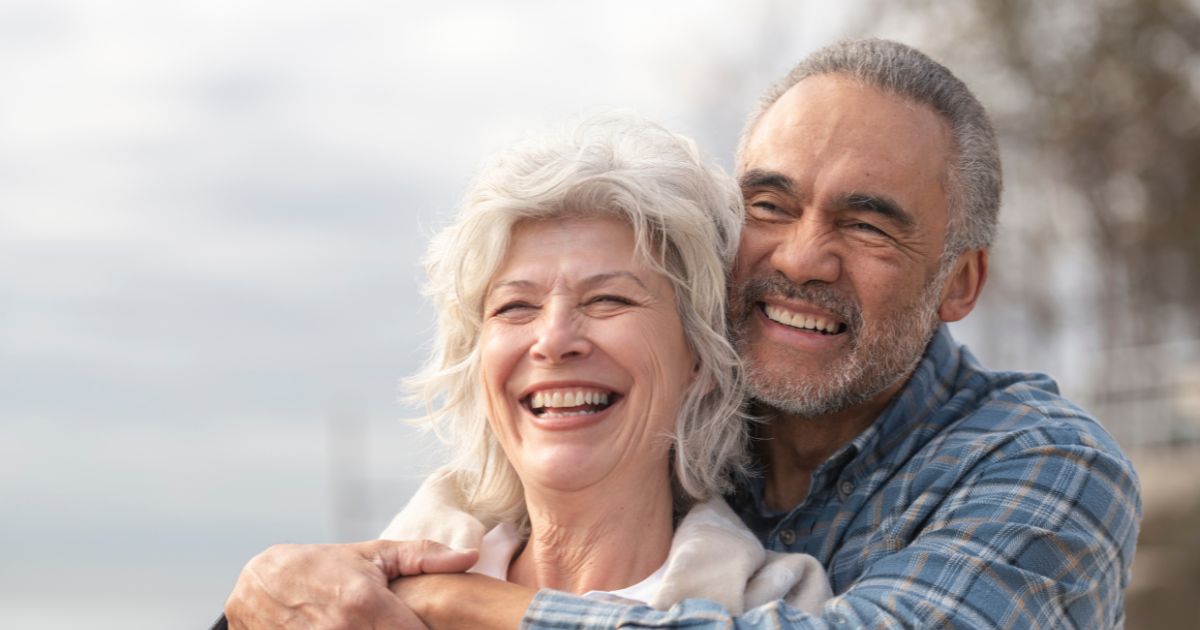 A smiling senior couple stands outdoors by the water, embracing and enjoying a joyful, peaceful moment together.