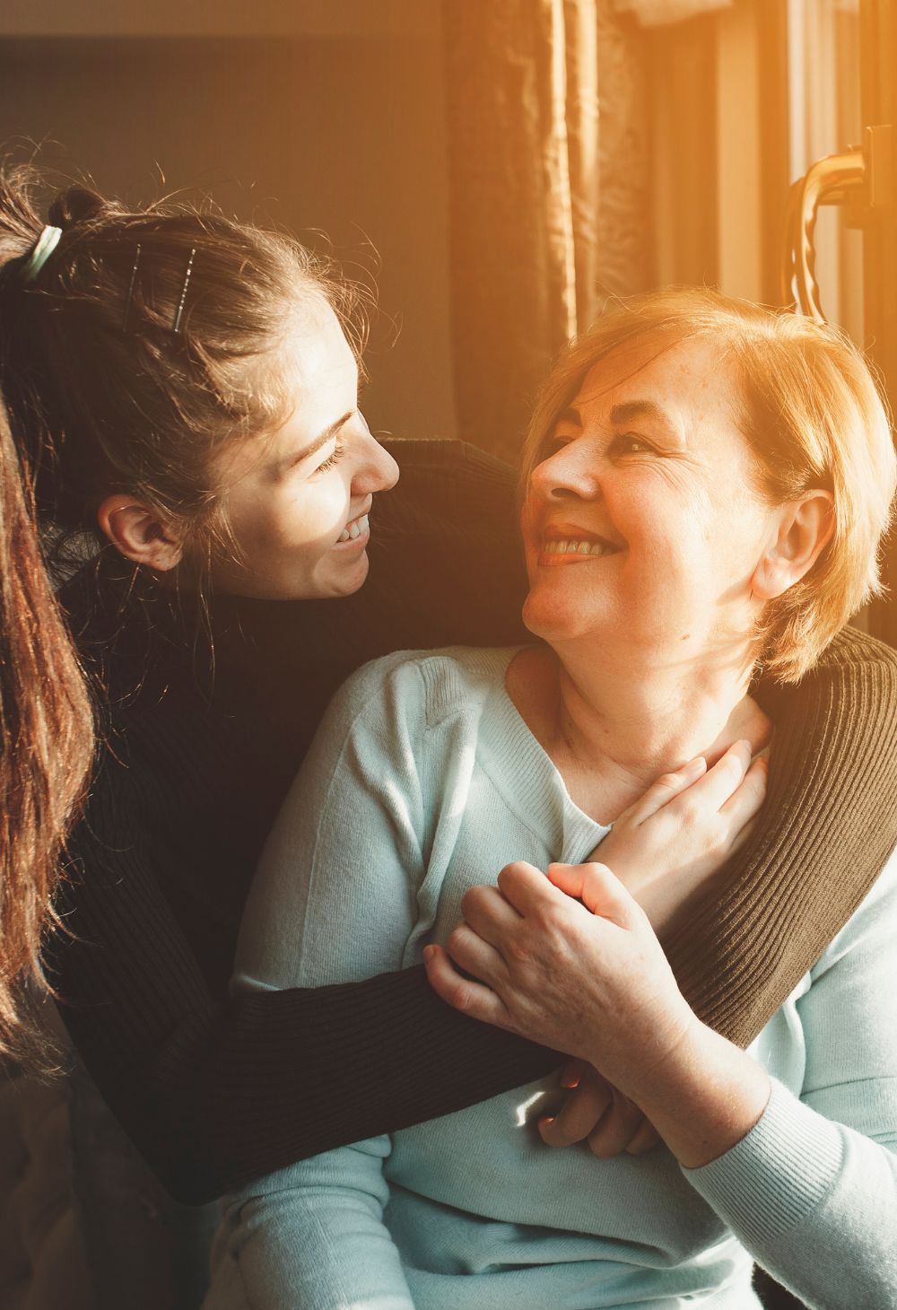 Compassionate Caregiver Sharing a Warm Embrace With Senior Woman at Home A young caregiver gently embraces a smiling senior woman while they sit together in a sunlit living room, sharing a moment of connection and comfort.