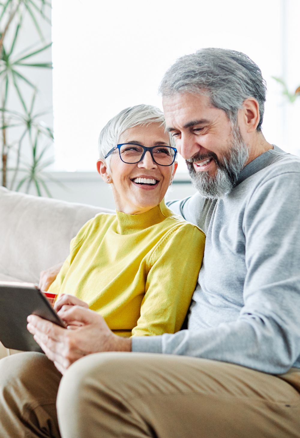 A smiling senior woman with short gray hair and glasses sits beside her partner on a couch while they look at a tablet together in a bright living room.