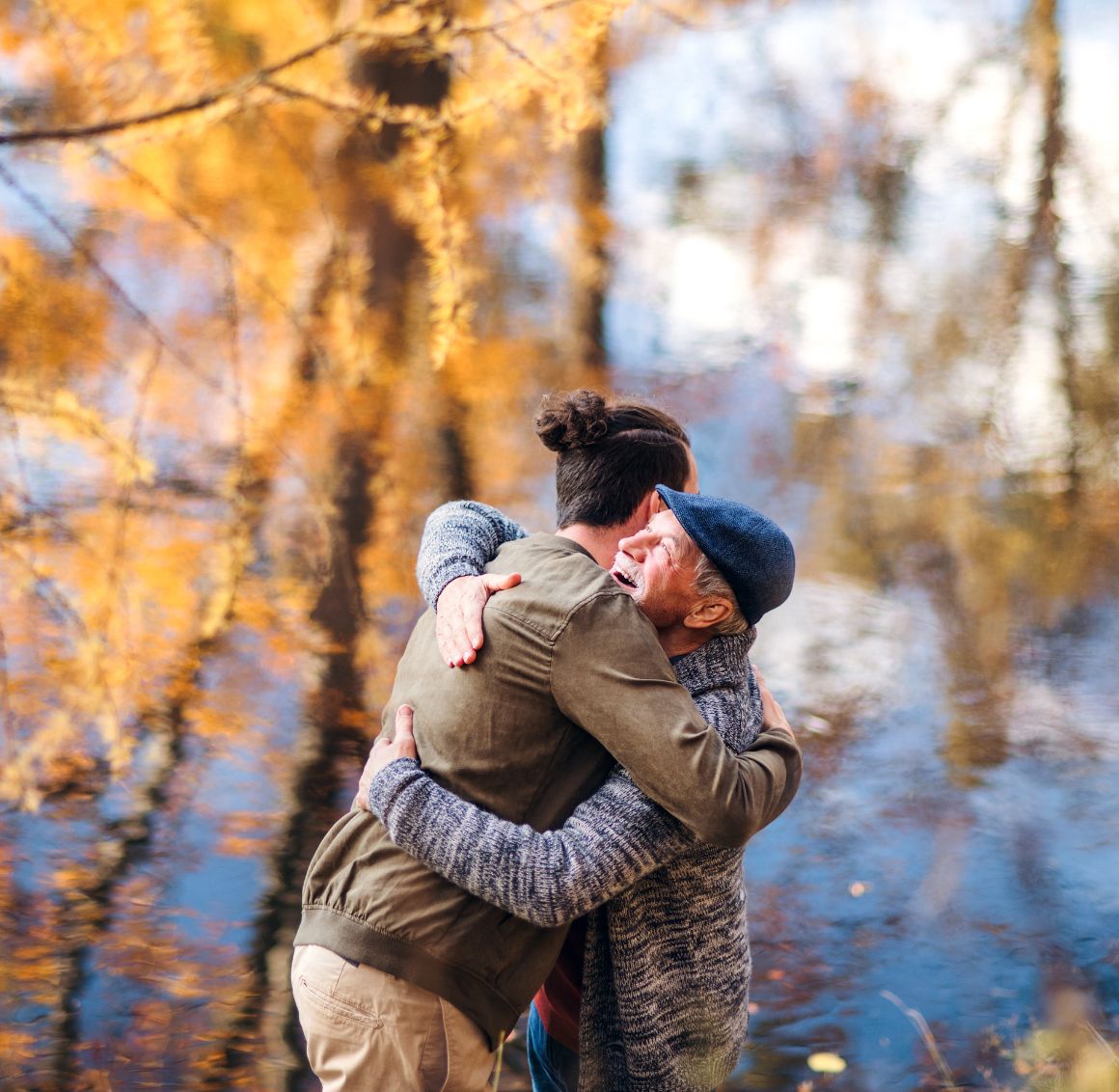 Senior man smiling while hugging a younger family member outdoors near a lake in autumn