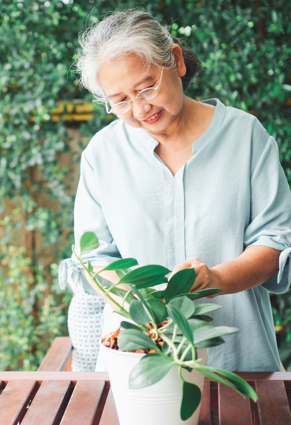 An elderly woman gently tends to a potted plant on a wooden table outdoors, representing peaceful gardening and independent senior living.