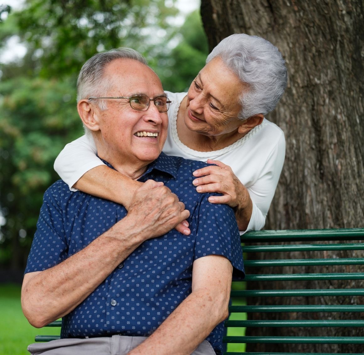 An elderly woman lovingly embraces her smiling partner from behind as they sit on a park bench. The couple looks happy and connected, enjoying a peaceful moment outdoors surrounded by greenery.