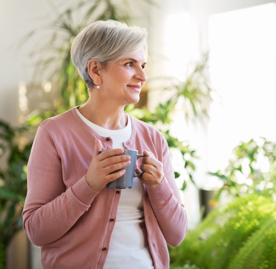 Smiling senior woman standing indoors holding a coffee mug in a bright, plant-filled living space