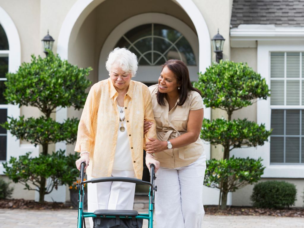 A professional caregiver supports an elderly woman using a walker outside a residential home surrounded by greenery.
