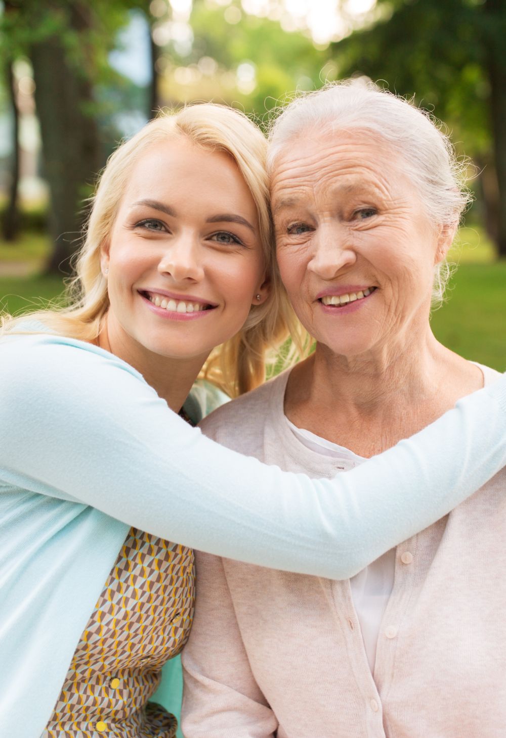 A smiling young woman warmly embraces her elderly mother as they sit together outdoors. Both women look happy and close, highlighting love, family connection, and emotional support in a peaceful park setting.