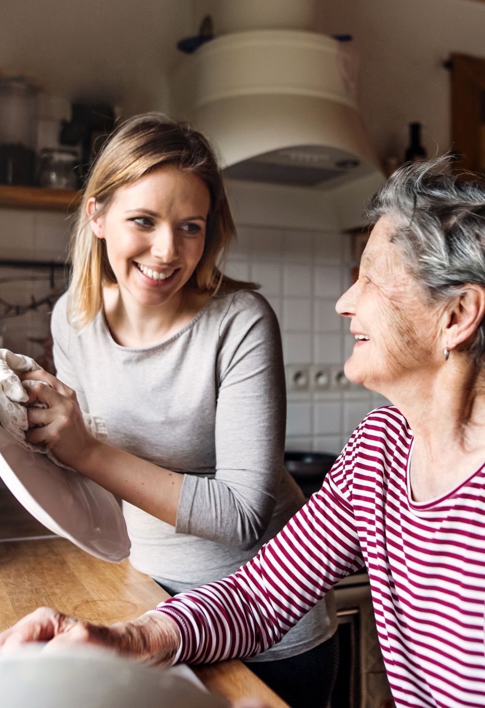 A smiling caregiver helps an elderly woman with dishes in a home kitchen, showing supportive and friendly in-home senior care.