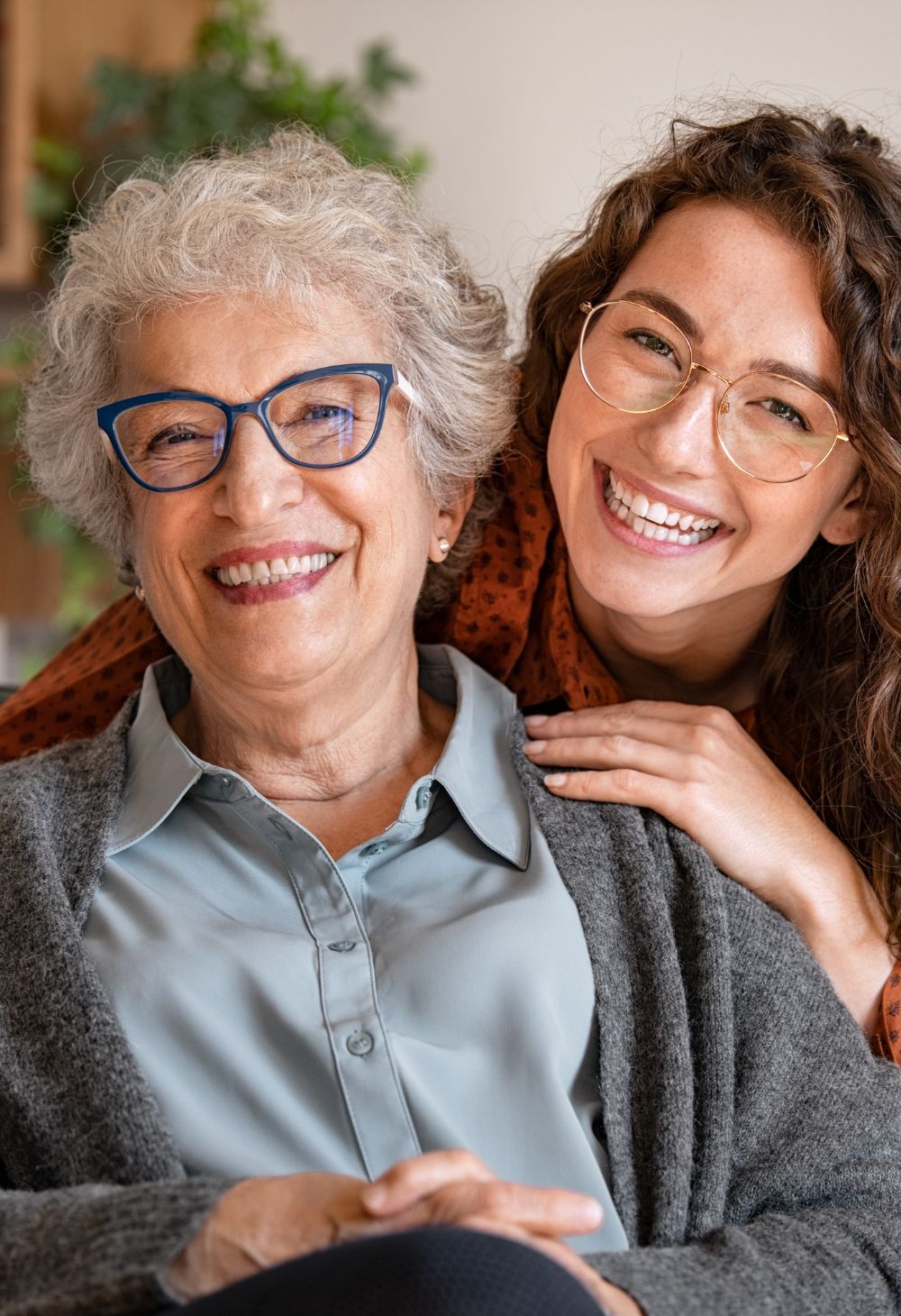 Smiling senior woman spending quality time with her adult daughter at home A joyful senior woman with short gray hair and blue glasses sits closely with her adult daughter, both smiling warmly at the camera. The daughter rests her hand gently on her mother’s shoulder, creating a loving and supportive family moment.