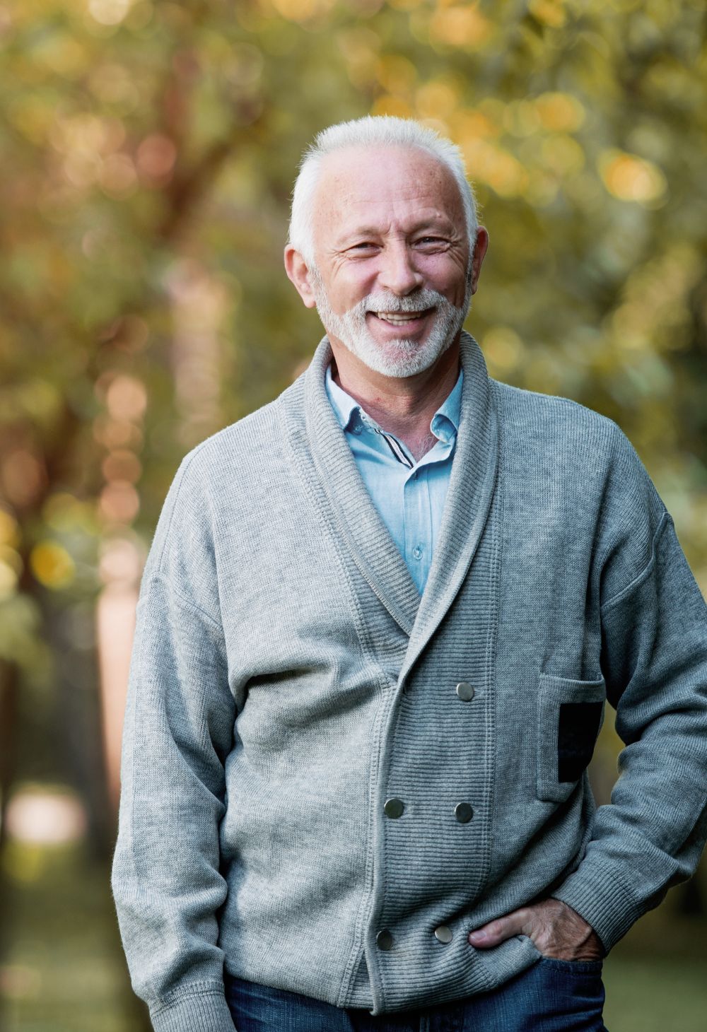 An older man with short white hair and a neatly trimmed beard smiles warmly while standing outside. He is wearing a light gray cardigan and blue shirt, with soft greenery in the background creating a calm, pleasant atmosphere.