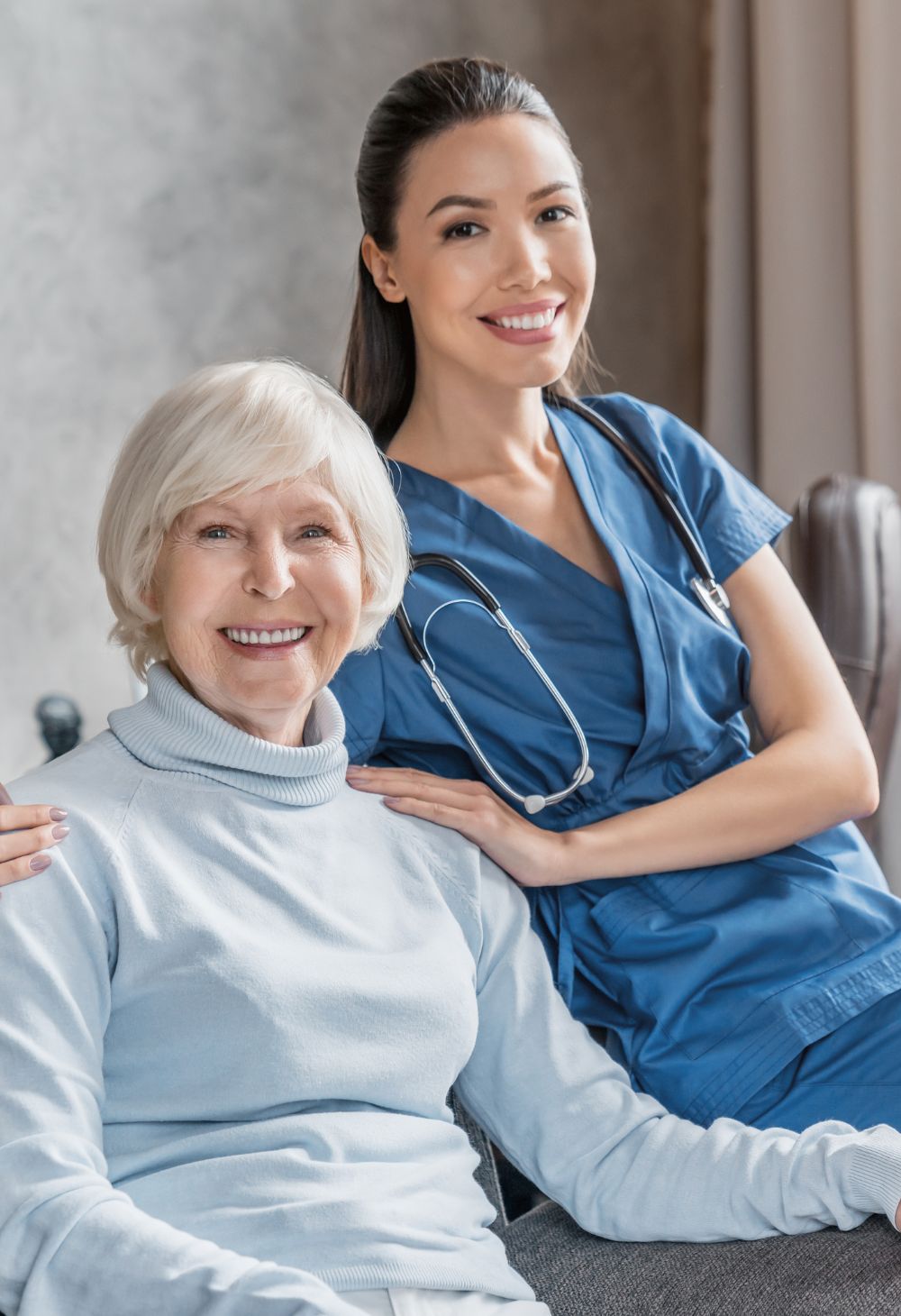 A senior woman sits and smiles warmly while a nurse in blue scrubs stands beside her with a hand on her shoulder. The scene highlights compassionate, supportive in-home care and a positive caregiver-client relationship.