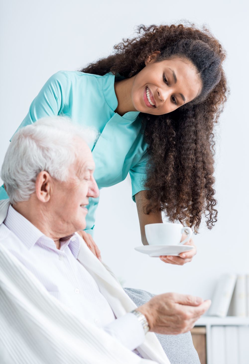 A smiling caregiver hands a cup of tea to an older man who is seated and wrapped in a blanket. The scene highlights gentle, compassionate assistance and supportive in-home care.