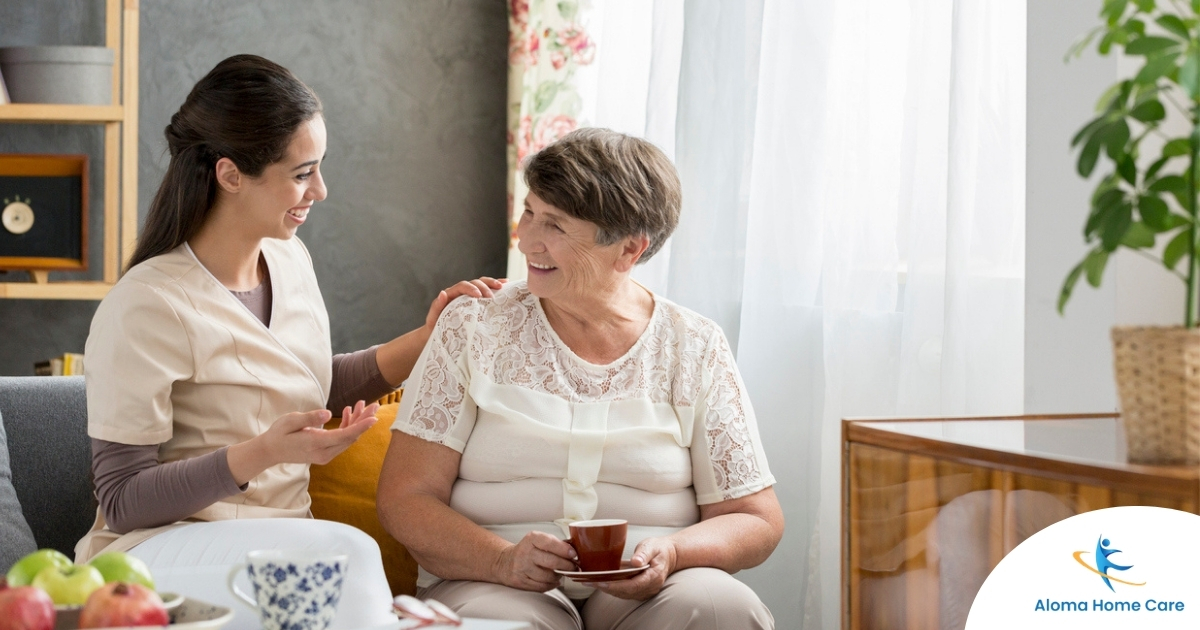 Smiling caregiver with her hand on an older woman’s shoulder as they share tea on the couch, showing home care vs assisted living